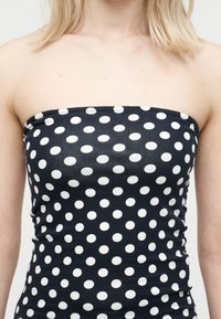 Γυναίκα με μαύρο τοπ χωρίς τιράντες με λευκές πουά, που δείχνει τους ώμους και το άνω μέρος του στήθους, σε απλό φόντο.