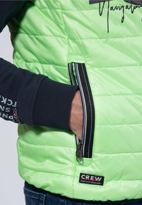 Jasnozielona kurtka puchowa z pikowaną teksturą, czarną kieszenią na suwak z paskowanymi akcentami i prążkowanym granatowym detalem na rękawie. Widoczna metka marki.