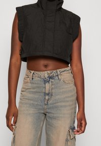 Svart kort väst med hög krage, tillverkad av texturerat material. Matchad med bleknande blå högmidjade jeans med en cargo-ficka.