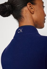 Tamsiai mėlyna palaidinė su aukštu apykakliu, su subtiliu išsiuvinėtu "CK" logotipu nugaroje. Lygus paviršius ir pritaikytas siluetas.