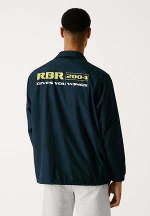 Navy blue windbreaker jacket with elastic cuffs, featuring “RBR 2004” and “GIVES YOU WINGS” printed in yellow and white on the back.