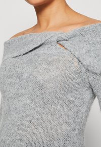 Gros plan d'une personne portant un pull gris en maille duveteuse à épaules dénudées avec un détail torsadé près de l'encolure.
