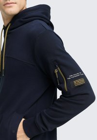 Marineblauwe hoodie gemaakt van zachte stof, met een ritszak op de mouw, gouden trekkoorden en een logo-opdruk op de arm.