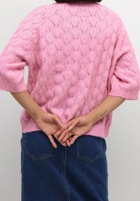 Rosa gestrickter Pullover mit Zopfmuster und kurzen Ärmeln, kombiniert mit einem Jeansrock. Die Hände sind hinten zusammengelegt, wodurch die Textur des Kleidungsstücks sichtbar wird.