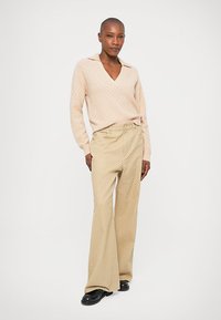 Femme debout portant un pull en V beige texturé et un pantalon large taille haute beige avec de petits motifs à pois et des chaussures noires.