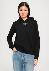 Jeune femme portant un sweat à capuche noir avec le texte "Just Cavalli True Lover" et un jean bleu clair, se tenant avec une main dans la poche devant un fond blanc.