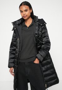 Casaco puffer preto com um design acolchoado brilhante, fecho completo, capuz destacado e bolsos com fecho, sobre uma camisola polo escura e jeans pretos.