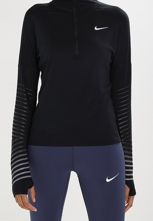 Kobieta ubrana w czarną, długorękawową bluzkę Nike z otworami na kciuki i niebieskie legginsy Nike, stojąca z ramionami swobodnie opadającymi wzdłuż ciała.