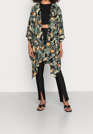 Femme portant un crop top noir et un pantalon avec des fentes à l'avant, des sandales noires et un kimono floral ample avec des motifs de feuilles vertes et orange.