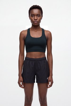 Une femme se tient face à l’objectif, vêtue d’un crop top de sport noir et d’un short de course, sur un fond blanc uni.