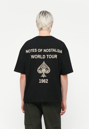 Zwarte katoen t-shirt met gouden tekst: "NOTES OF NOSTALGIA WORLD TOUR 1962," met een decoratief schoppenontwerp op de achterkant. Korte mouwen.
