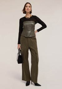 Top in pelle verde oliva a corsetto abbinato a pantaloni cargo larghi verde oliva. Top lungo trasparente nero a maniche lunghe e tacchi a stiletto neri. Piccola borsa nera.