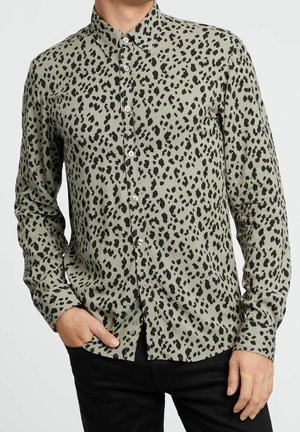 Homme portant une chemise à manches longues boutonnée avec un imprimé léopard vert et noir, associée à un pantalon noir, main dans la poche.