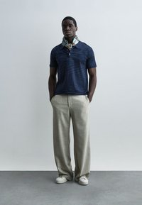 Άνδρας που φοράει μπλε σκούρο πόλο πουκάμισο, μπεζ παντελόνι με φαρδιά μπατζάκια, λευκά αθλητικά παπούτσια και μαντήλι με σχέδια, στέκεται με τα χέρια στις τσέπες μπροστά σε απλό φόντο.