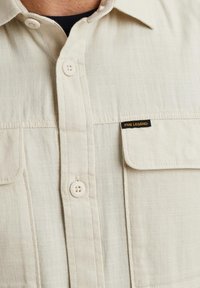 Helles beiges Hemd mit Knopfleiste aus strukturiertem Stoff, ausgestattet mit zwei Brusttaschen und einem kleinen schwarzen Logo-Patch, mit Kunststoffknöpfen.