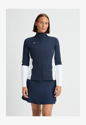 Marineblau und weiße Zip-Up Sportjacke mit langen Ärmeln und hohem Kragen, kombiniert mit einem marineblauen Rock. Glatter, dehnbarer Stoff, schlankes Design.