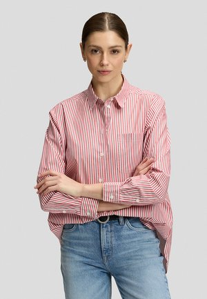Donna con capelli castani raccolti in uno chignon basso indossa una camicia a righe rosse e bianche e jeans blu, con le braccia incrociate, in piedi davanti a uno sfondo neutro.