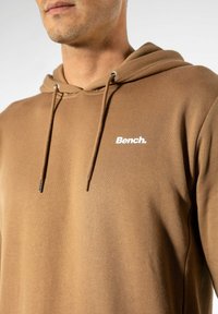 Brauner Hoodie mit weichem Material, ausgestattet mit einer Kängurutasche und einer Kapuze mit Kordelzug. Das Logo "Bench." ist in Weiß auf der Brust aufgedruckt.