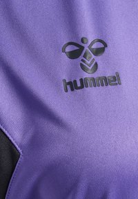 Camisa deportiva morada con tejido texturizado, logo negro de "hummel" y un elemento de diseño abstracto en la parte frontal.