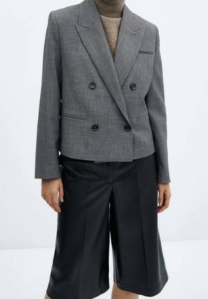 Personne portant un blazer gris croisé sur un col roulé beige, associé à un short noir ample arrivant aux genoux.