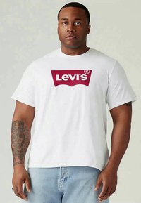 Homem com o braço esquerdo tatuado, vestido com uma t-shirt branca da Levi's e jeans azul claro, em pé contra um fundo claro.