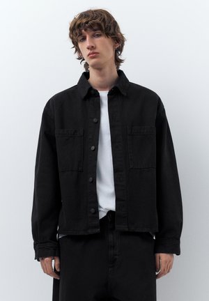 Mladý človek s kučeravými vlasmi, oblečený v čiernej zapínacej bunde cez biele tričko, stojaci pred jednoliatym svetlým pozadím.