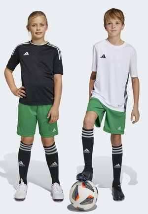 Due bambini in pantaloni da calcio verdi, uno indossa una maglietta nera, l'altro è in bianco. Entrambi indossano calze a righe nere e stanno in piedi con un pallone.