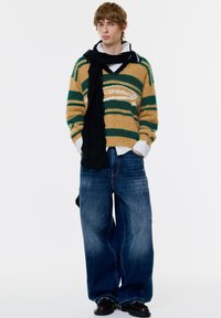 Hnedo-zelený pruhovaný chlpatý sveter s nápisom "COMMUNITY", skombinovaný s širokými tmavomodrými džínsami a čiernym šálom, nosený cez bielu košeľu.