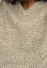 Pletený béžový svetr s výstřihem do V, jemnou texturou a žebrovaným lemem. Materiál vykazuje lehké barevné variace a jemnou nadýchanost.