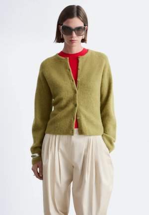 Kvinde iført store mørke solbriller, olivengrøn knapstrikket cardigan over rød top og løse beige plisserede bukser, stående mod en hvid baggrund.