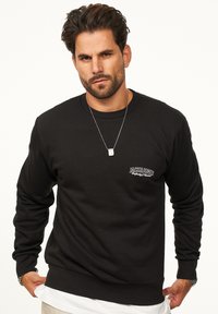 Black sweatshirt with ribbed texture, crew neck, and a small white logo on the left chest. Model wears a silver necklace underneath.