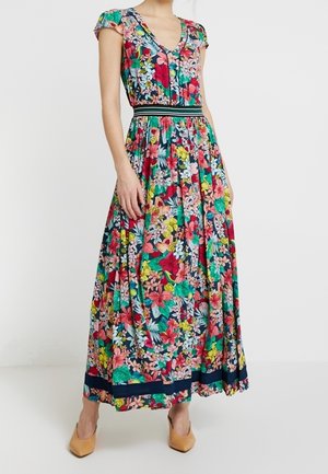 Femme portant une robe maxi florale colorée avec des manches courtes ballon, un col en V, une ceinture rayée noir et blanc et des talons couleur beige.