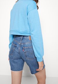 Cropped blue sweatshirt with long sleeves paired with high-waisted denim shorts featuring frayed hems and back pockets with a logo patch.