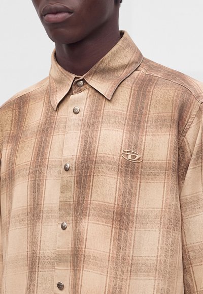 Homme portant une chemise à carreaux beige et marron boutonnée avec col et petit logo ovale sur la poitrine.
