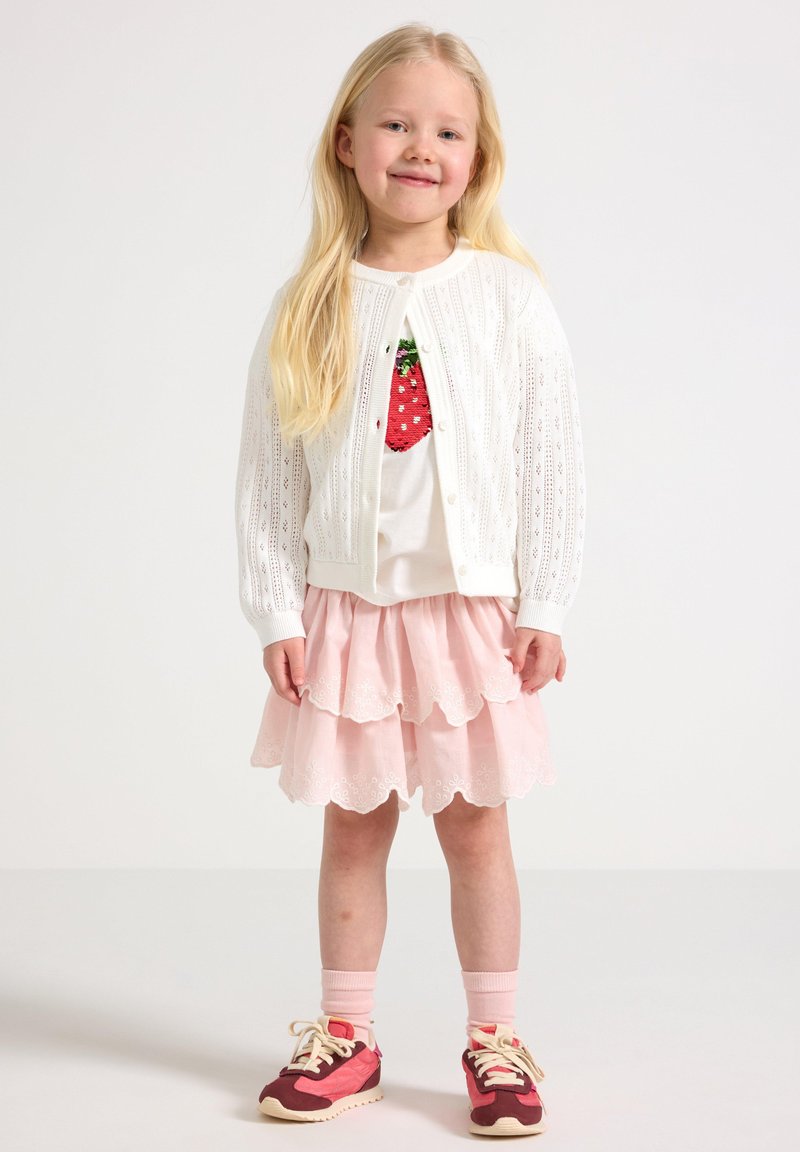 Cardigan bianco lavorato a maglia con motivo a occhio di pernice, camicia bianca con grafica di fragole, gonna rosa a strati e sneaker rosse con dettagli in crema.