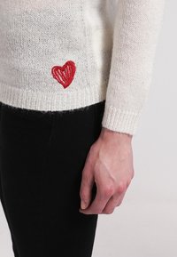 Pull en maille blanche avec un petit cœur rouge brodé près de la taille, porté avec un pantalon noir, montrant la main droite de la personne détendue le long du corps.