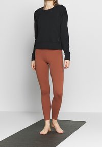 Sudadera negra de manga larga y cuello redondo, combinada con leggings marrones ajustados, ambos de un tejido suave sobre un tapete gris.