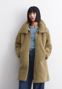 Světlý, nadýchaný, oversized kabát s jemnou texturou a velkým límcem. Obsahuje celý zip a dvě boční kapsy, nošený přes modrý top.