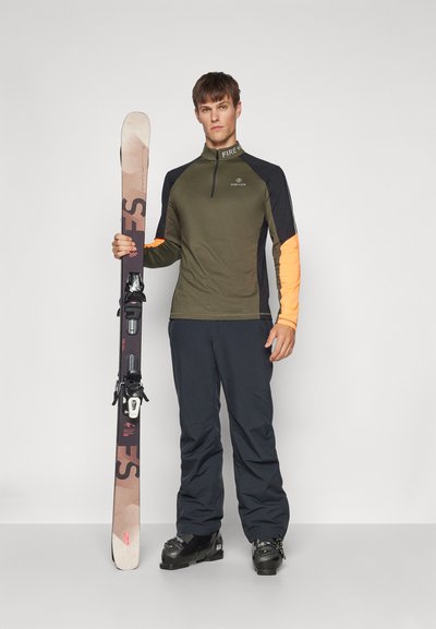 Ski outfit featuring an olive green long-sleeve top with orange accents, dark pants, and black ski boots, holding brown skis with bindings.