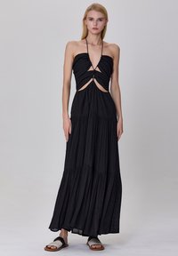 Robe maxi noire avec un décolleté en V profond, des découpes, une jupe en couches et un tissu léger. Associée à des sandales plates.