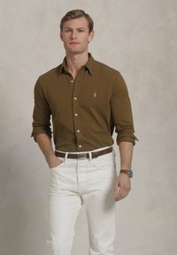 Rjava srajca z dolgimi rokavi in logotipom na prsih, v kombinaciji z belimi kavbojkami in rjavim pasom. Na zapestju je ura.