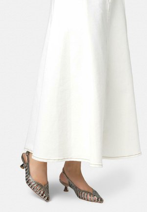 Pies con zapatos de tacón pequeño en bloque, de malla metálica y estilo slingback, combinados con una falda blanca hasta el tobillo.