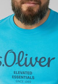 Turkio spalvos trumpas rankovėmis marškinėliai, pagaminti iš minkštos medžiagos, su juodu tekstu, skaitančiu "s.Oliver Elevated Essentials Since 1969."