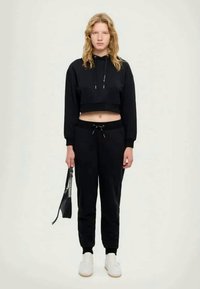 Black cropped hoodie with drawstrings, paired with black sweatpants. Model holds a small black bag. White sneakers complete the outfit.