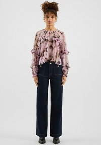 Przezroczysta różowa bluzka z abstrakcyjnymi brązowymi wzorami, z falbanami na rękawach, w zestawieniu z ciemnymi, szerokimi jeansami i czarnymi botkami za kostkę.