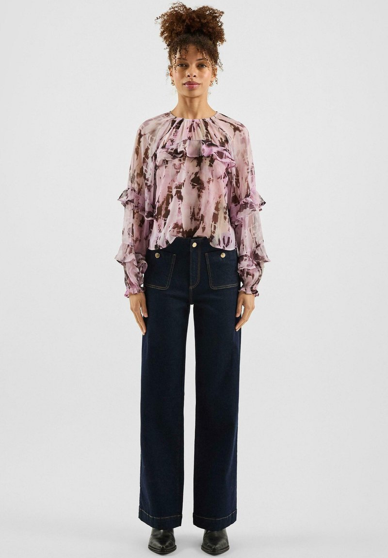 Przezroczysta różowa bluzka z abstrakcyjnymi brązowymi wzorami, z falbanami na rękawach, w zestawieniu z ciemnymi, szerokimi jeansami i czarnymi botkami za kostkę.