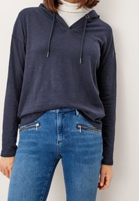 Tmavě námořnická mikina s výstřihem do V, šňůrkami a dlouhými rukávy, doplněná světle modrými džíny s kapsami na zip na přední straně.