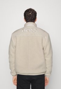 Beige shearlingjacka med hög krage, med en kontrasterande ljusbrun panel på axlarna broderad med "BOSS" i vitt.