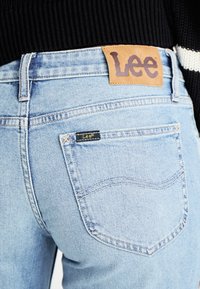 Light blue denim jeans with a leather Lee label on the waistband, featuring a back pocket with a small Lee tag and stitched detailing.