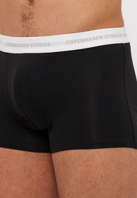 Schwarze Boxershorts aus glattem Stoff mit einem weißen elastischen Bund, der das Logo "Copenhagen Studios" in grauer Schrift zeigt.